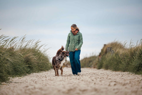 Eine Frau läuft mit ihrem Hund neben sich an der Leine durch einen Sandweg in den Dünen. Sie trägt eine grüne Jacke und sieht den Hund dabei an.
