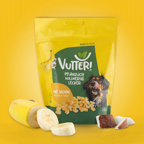 Neben einer Tüte veganen Leckerli wie Huhn von Vutter! liegen Bananenstücke und Kokosnuss