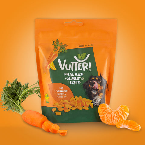 Neben einer Tüte veganen Leckerli wie Leerwurst von Vutter! liegt eine Karotte und ein Mandarinen-Stücke